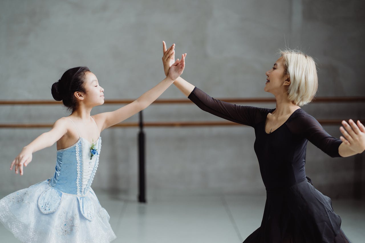 Side view of graceful Asian girl wearing tutu standing near teacher in black outfit with raised arms in ballet studio
