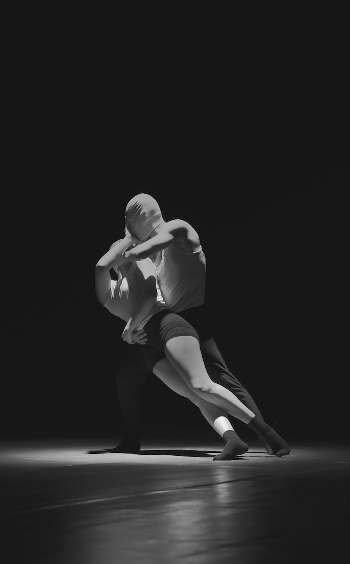 Two contemporary dancers perform an intense masked routine in a dark studio setting.
